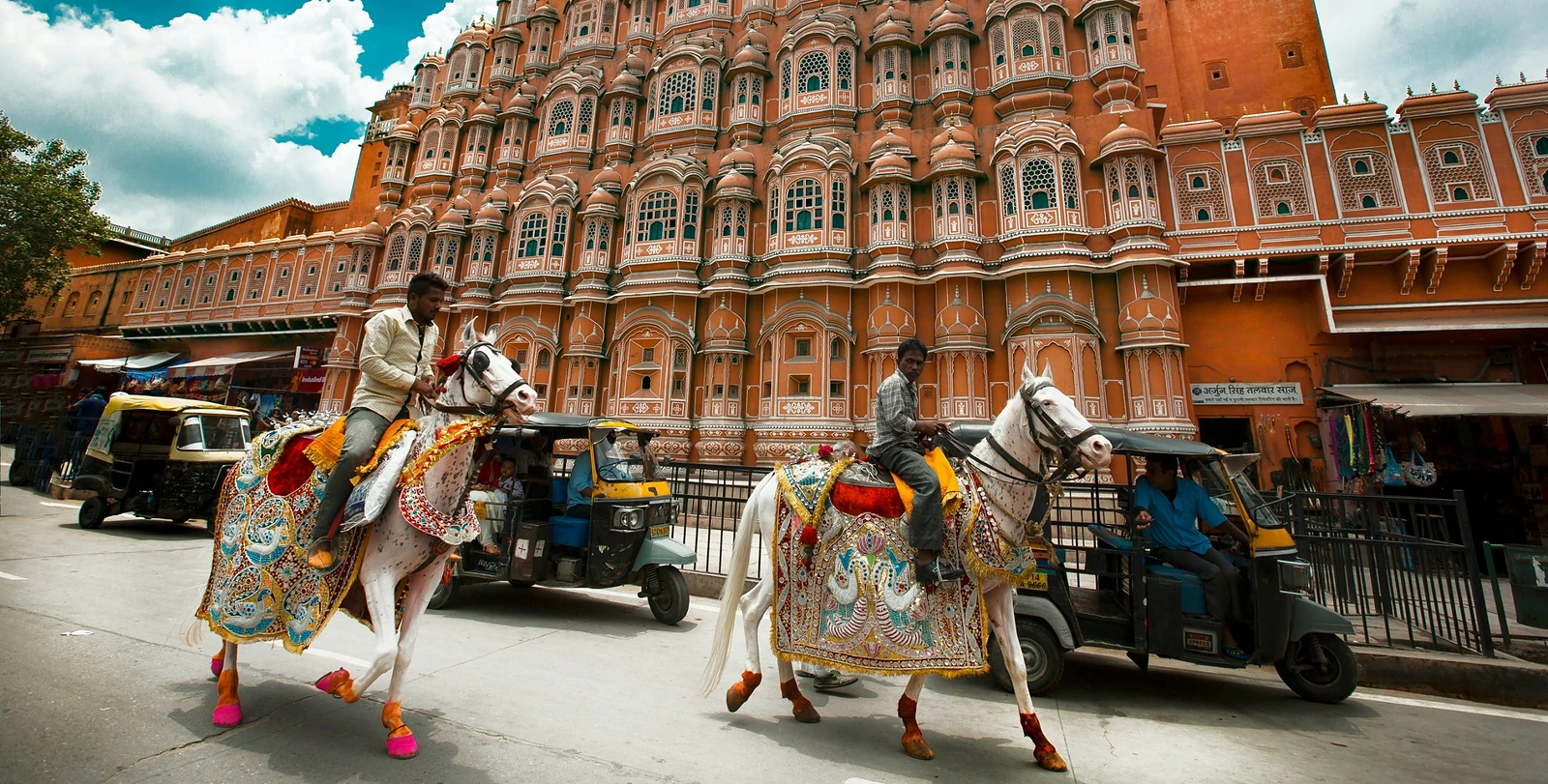Dois cavaleiros em cavalos brancos enfeitados passam diante do Hawa Mahal de arenito rosa, em Jaipur