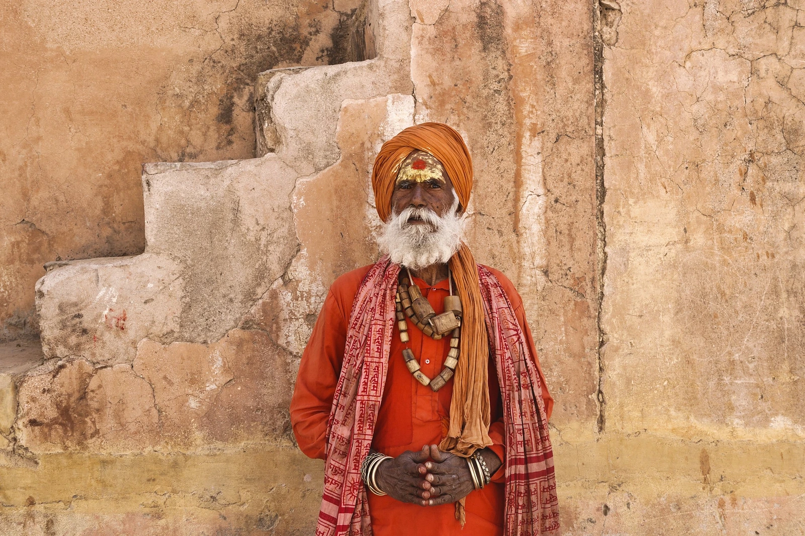 An elderly sadhu in a saffron turban and robe, with a shawl printed with the name of Ram, standing against an old ochre wall